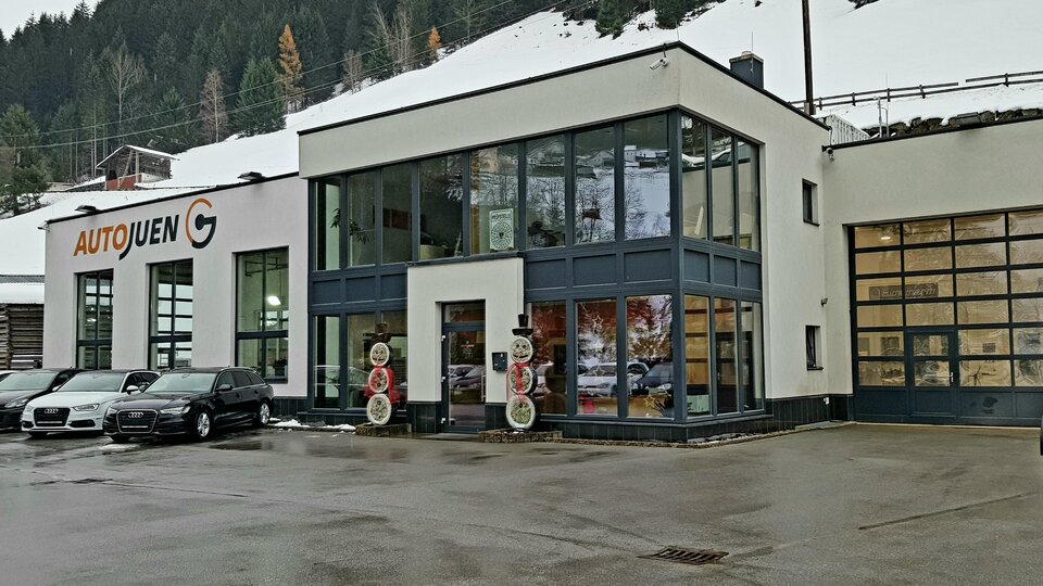 A modern car dealership with large windows and contemporary architecture. Several vehicles are parked in front of the building, and snow-capped mountains can be seen in the background.
