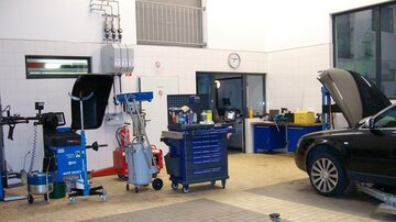A workshop with various tools and equipment. In the foreground, there is a black vehicle with the hood open.