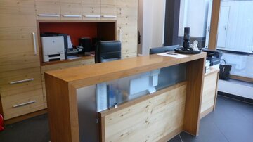 A modern reception area with wood paneling and a desk. In the background, office equipment and a large window area can be seen.