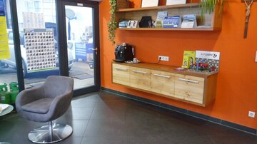 A modern reception area with an orange wall, a wooden shelf, and a comfortable gray armchair. On the shelf, there are informational materials and a coffee machine.