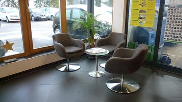 A modern waiting room with three gray armchairs and a small table. Through large windows, cars and a green plant are visible.