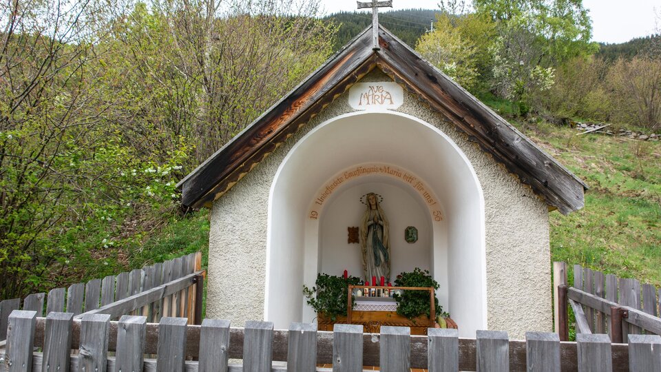 A small chapel with a holy statue stands in nature. It is surrounded by a wooden fence and enclosed by trees. | © Bernhard Gruber – Photography