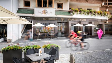 A bakery with an elegant outdoor area and sun umbrellas. Two cyclists are riding along the street.