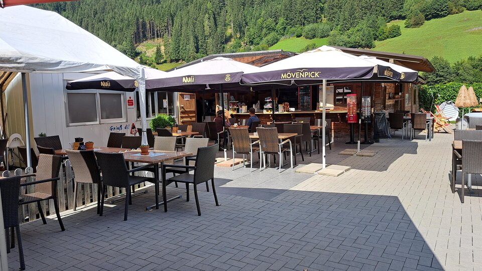 A cozy outdoor area of a restaurant with tables and chairs. In the background, there is the restaurant with umbrellas and a green landscape.