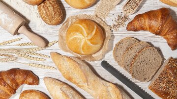 A selection of fresh bread, rolls, and croissants is laid out on a light wooden surface. Wheat grains and a rolling pin are also visible. | © MPREIS Warenvertriebs GmbH