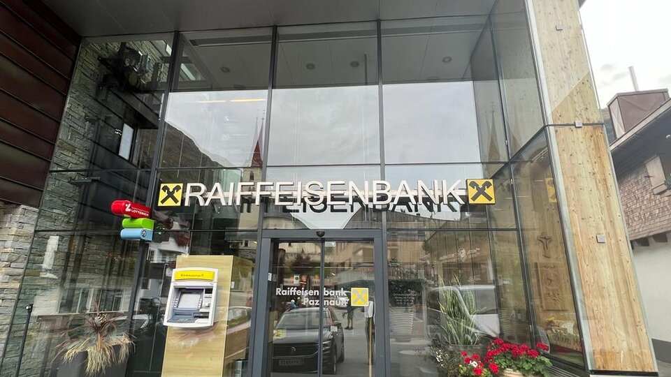 Eingang zur Raiffeisenbank mit moderner Architektur. An der Fassade sind ein Geldautomat und Pflanzen zu sehen.