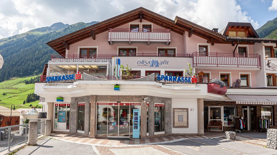 A cozy building in the mountains with a clear sky. There are shops and balconies with colorful flowers.