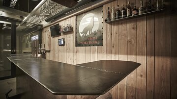 A stylish bar with a wooden design and a metal counter. Shelves with spirits and wall decorations give the space an inviting atmosphere.
