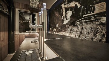 A modern bar with a long counter and taps. In the background, there is a window featuring a picture of construction machinery and mining.