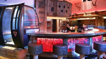 A modern bar with a curved counter and stylish bar stools. The interior features wood and has vibrant colored lighting.