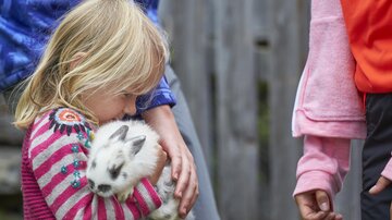 The image shows a blonde child holding a white-gray spotted rabbit tightly and carefully in their arms. The child is wearing a colorful patterned sweater and gently leans their head down towards the animal, making the moment appear very calm and affectionate.

On the right side of the image, another child is only partially visible, while adults or older individuals stand blurred in the background. The focus is clearly on the closeness between the child and the rabbit. The scene conveys a sense of security, care, and childlike affection for animals.