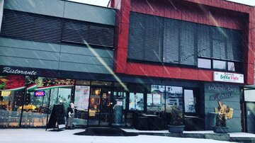 A modern restaurant building with a red and gray facade in the snow. In front of the entrance, some chairs and tables as well as decorations are visible.