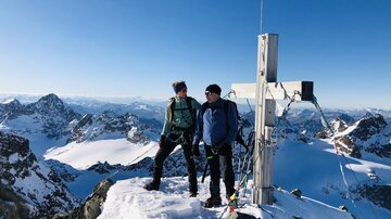 Two climbers are standing on a snow-covered summit. In the background, impressive mountains and a clear blue sky can be seen.