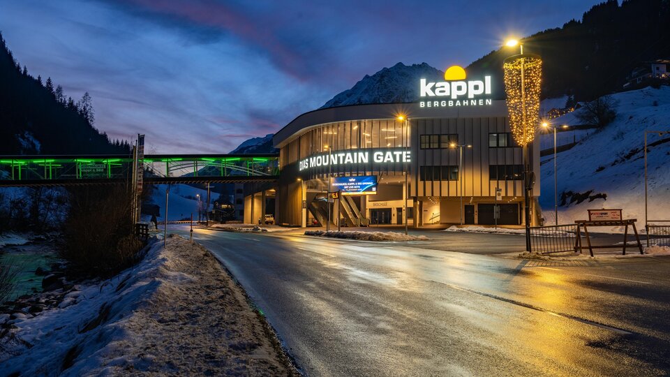 A modern building with the inscription "Kappl Bergbahnen" stands in the foreground. The scene shows a wintry environment at dusk.