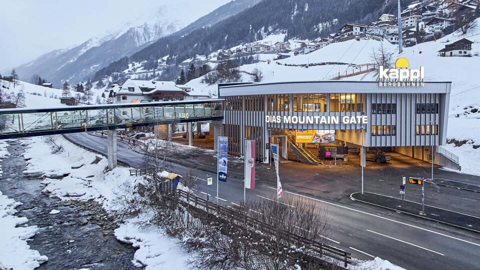 A modern building with the inscription "THE MOUNTAIN GATE" stands in a snow-covered landscape. In the background, mountains and a river are visible.