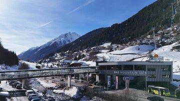 A snow-covered valley with high mountains in the background. In the foreground, there is a modern building with a bridge and parking lots.