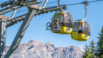 A cable car with yellow gondolas travels over the trees. In the background, majestic mountains and a blue sky can be seen.