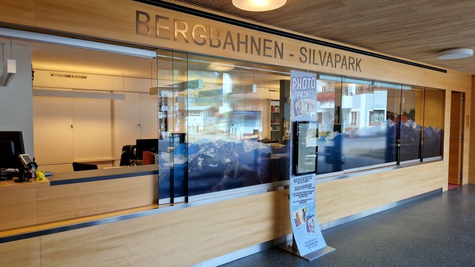 Ein Empfangsbereich mit einer Holzverkleidung und einem großen Glasfenster. An der Wand steht „BERGBAHNEN - SILVAPARK“.