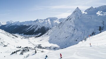 Eine schneebedeckte Berglandschaft mit hohen Gipfeln und klarem blauen Himmel. Zwei Skifahrer fahren die Piste hinunter.