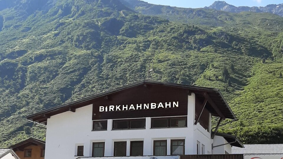 Eine Bergbahnstation mit dem Namen "Birkhahnbahn" befindet sich im Vordergrund. Im Hintergrund sind grüne Berge und ein blauer Himmel zu sehen.