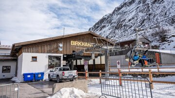 Ein modernes Skigebäude mit dem Namen "Birkhahn" und einem Sessellift im Hintergrund. Die Umgebung ist schneebedeckt und von Bergen umgeben.