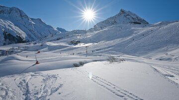 Eine verschneite Berglandschaft unter klarem, blauem Himmel. Die Sonne strahlt hell über die schneebedeckten Hügel.