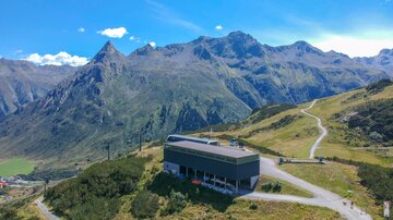 Eine moderne Hütte in den Bergen mit umliegenden Bergen und einem klaren blauen Himmel. Der Weg führt durch die grüne Landschaft in Richtung der Berge.