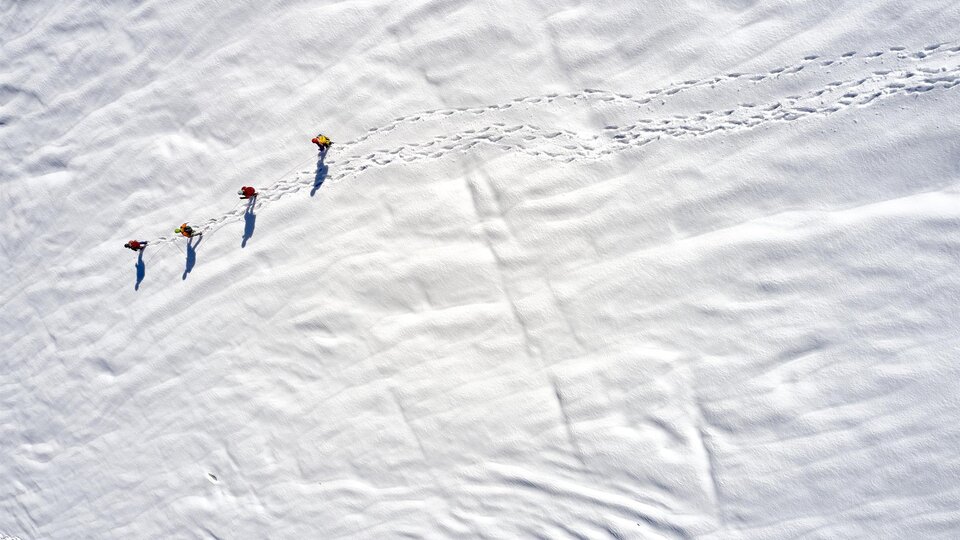 Four people are walking across a snow-covered area. Their tracks are clearly visible in the fresh snow.