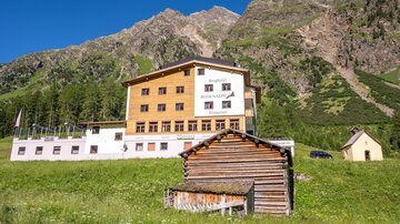Ein modernes Gebäude in einer bergigen Landschaft. Im Vordergrund steht ein traditionelles Holzhaus.