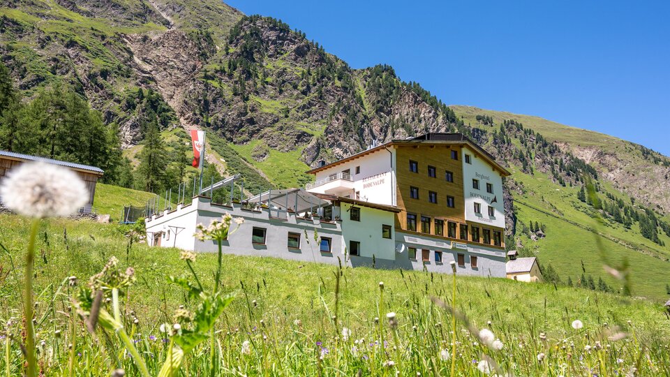 Ein großes Gebäude in den Bergen, umgeben von grünen Wiesen und Felsen. Der Himmel ist klar und blau.
