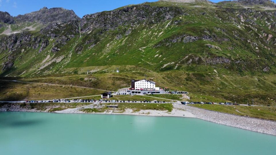 Ein malerischer Bergsee mit hellem, türkisfarbenem Wasser. Am Ufer steht ein großes, weißes Gebäude und grünes Gebirge umgibt die Landschaft.