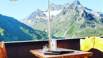 Ein gemütlicher Tisch mit roten Sitzkissen unter einem Sonnenschirm. Im Hintergrund erstrecken sich beeindruckende Berge und ein blauer Himmel.