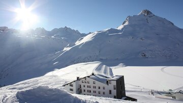 Eine malerische winterliche Landschaft mit schneebedeckten Bergen. Im Vordergrund befindet sich ein modernes Hotel, umgeben von verschneiten Pisten.
