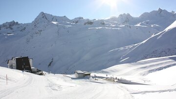 Eine schneebedeckte Berglandschaft mit strahlendem Sonnenschein. Im Vordergrund sind Gebirgshütten und Skipisten zu sehen.