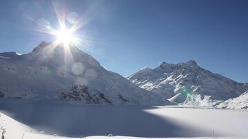 Eine winterliche Berglandschaft mit schneebedeckten Gipfeln und strahlendem Sonnenlicht. Der Himmel ist klar und blau.
