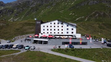 Ein modernes Hotel in den Bergen mit einer Terrasse und einem Parkplatz. Umgeben von grünen Hügeln und einer malerischen Landschaft.