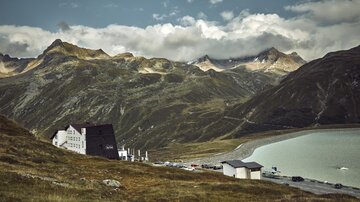 Eine malerische Berglandschaft mit einem See im Vordergrund. Im Hintergrund sind majestätische Berge und eine charmante Unterkunft zu sehen.