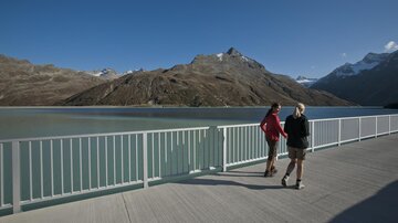 Zwei Personen spazieren entlang einer Uferpromenade mit Blick auf einen ruhigen See und majestätische Berge. Der Himmel ist klar und sonnig.