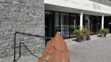 Ein moderner Berggasthof mit einem Eingangsbereich und einem stilisierten Schild der Bike Arena. Die Umgebung ist mit Pflanzen und einem klaren Himmel geschmückt.