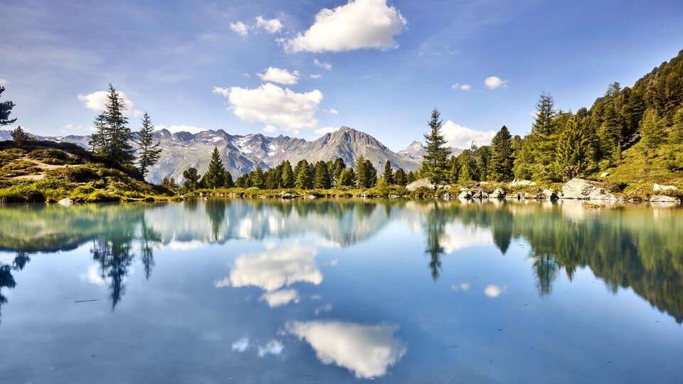 A tranquil mountain lake with crystal-clear water and reflecting clouds. The surrounding trees and mountains create an idyllic natural scene.