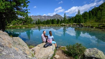 A tranquil lake surrounded by mountains and trees. Two people are sitting on the shore and enjoying the view.