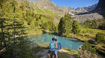 A couple stands by a clear lake in a beautiful mountain landscape. The surrounding mountains are green and sunny.