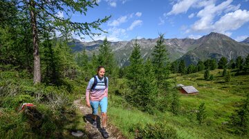 A woman hikes along a path through a green landscape. In the background, mountains and a small cabin can be seen.