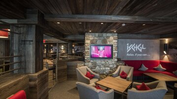An elegant restaurant with cozy seating areas and a warm wooden ceiling. A television is mounted on the wall, integrated into the modern, welcoming atmosphere.