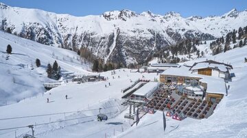 A beautiful winter landscape with snow-covered mountains. In the foreground, there is a ski chalet and lift facilities, surrounded by skiers.