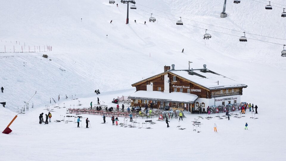A wooden house in the mountains, surrounded by snow and skiers. In the background, ski lifts can be seen.