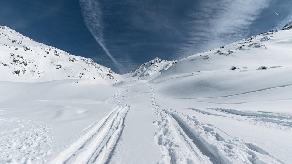 A snowy landscape with tracks in the snow and high mountains. The sky is blue with some clouds.