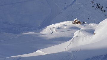 A snow-covered landscape with gentle hills and a small cabin. Clear blue skies and snow-capped mountains create a tranquil atmosphere.