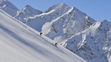 Two skiers are descending a snow-covered slope. In the background, impressive, snow-covered mountains can be seen.