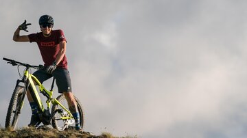 Ein Radfahrer posiert mit seinem Mountainbike auf einem Hügel. Im Hintergrund sind Wolken am Himmel zu sehen.